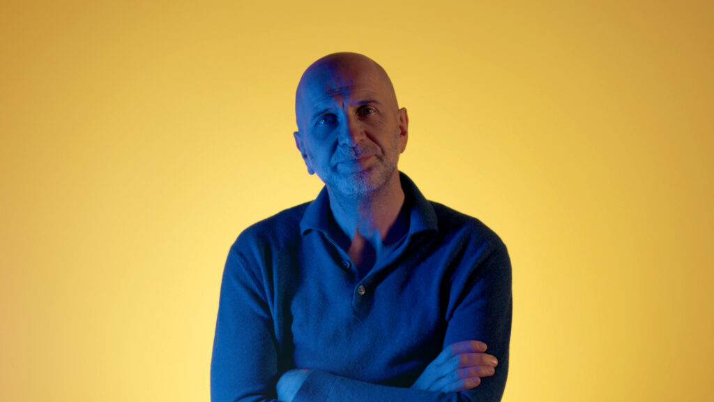Nic Balthazar die voor een gele achtergrond met gekruiste armen poseert voor De UGent